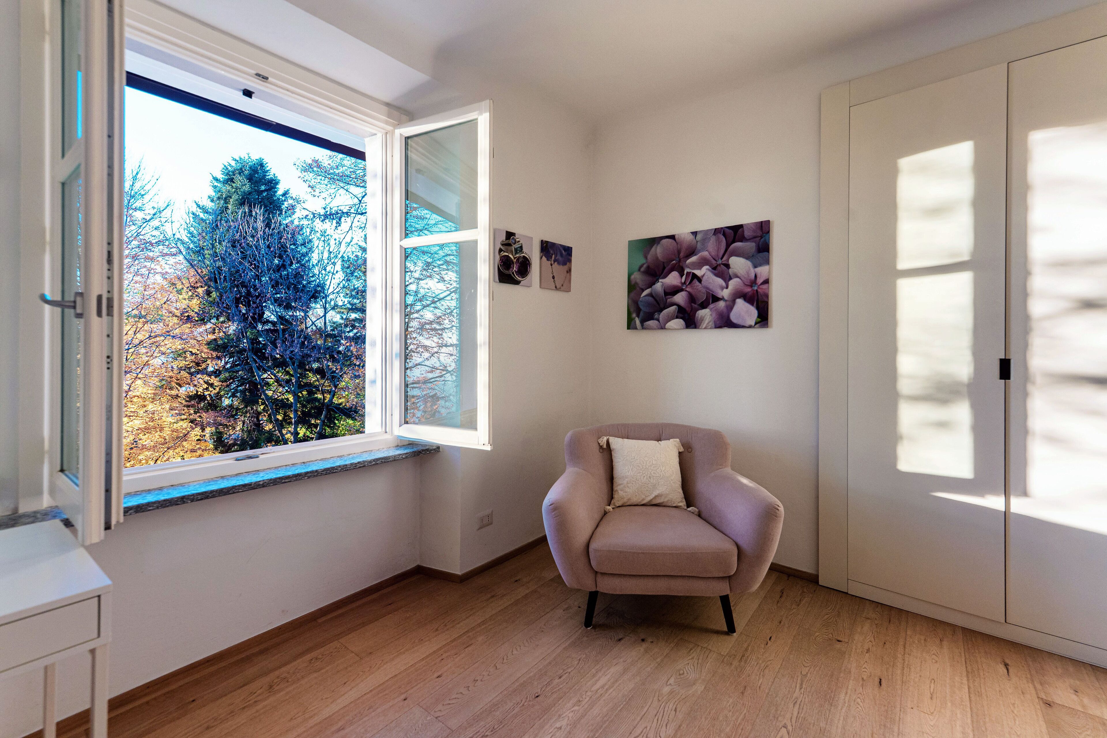 A room with a wooden floor, a beige armchair, a window with a view of trees, and a door.
