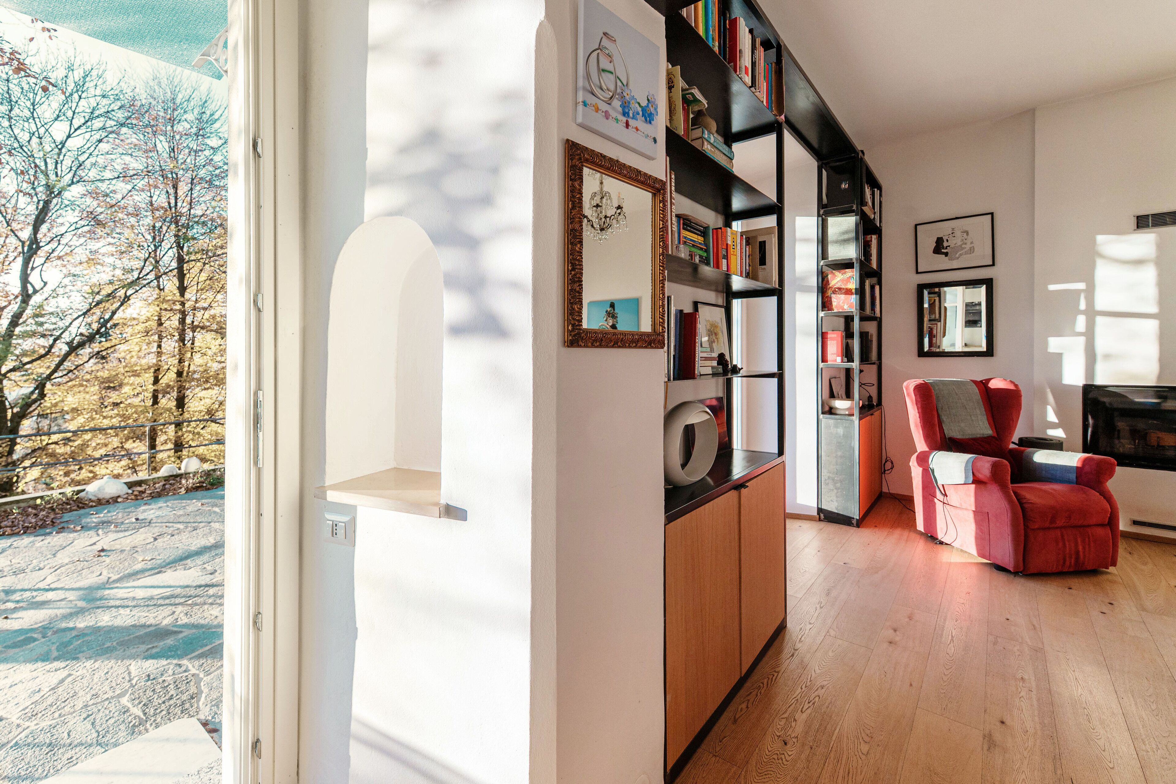 Un salon moderne avec un plancher en bois, un fauteuil rouge, une bibliothèque et une grande fenêtre donnant sur un paysage enneigé.
