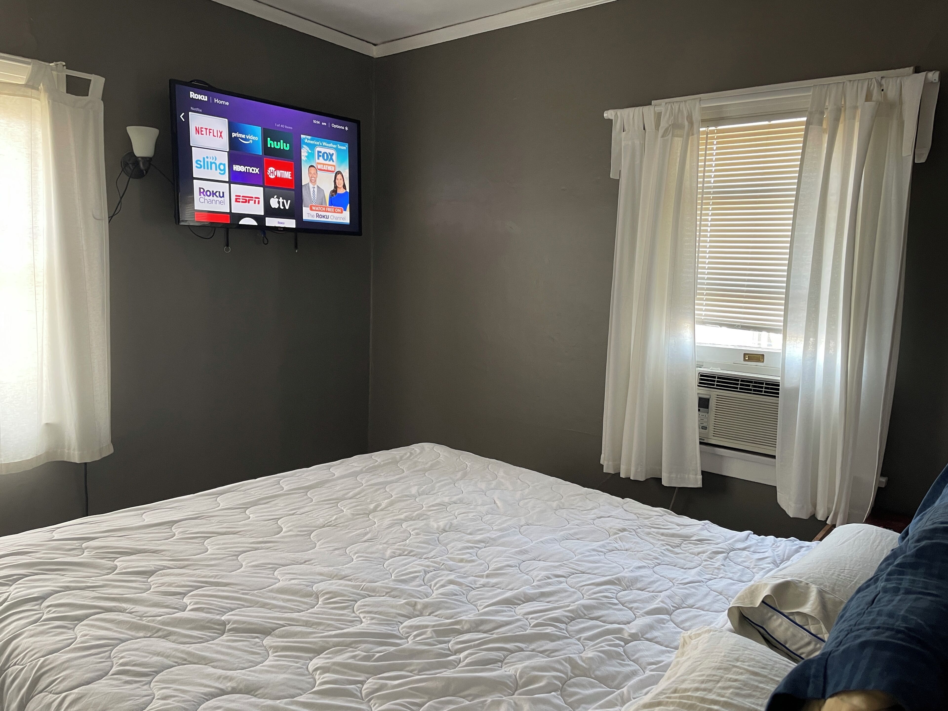 Een hotelkamer met een bed, een aan de muur bevestigde televisie waarop verschillende streamingdiensten worden getoond, en een raam met gordijnen.