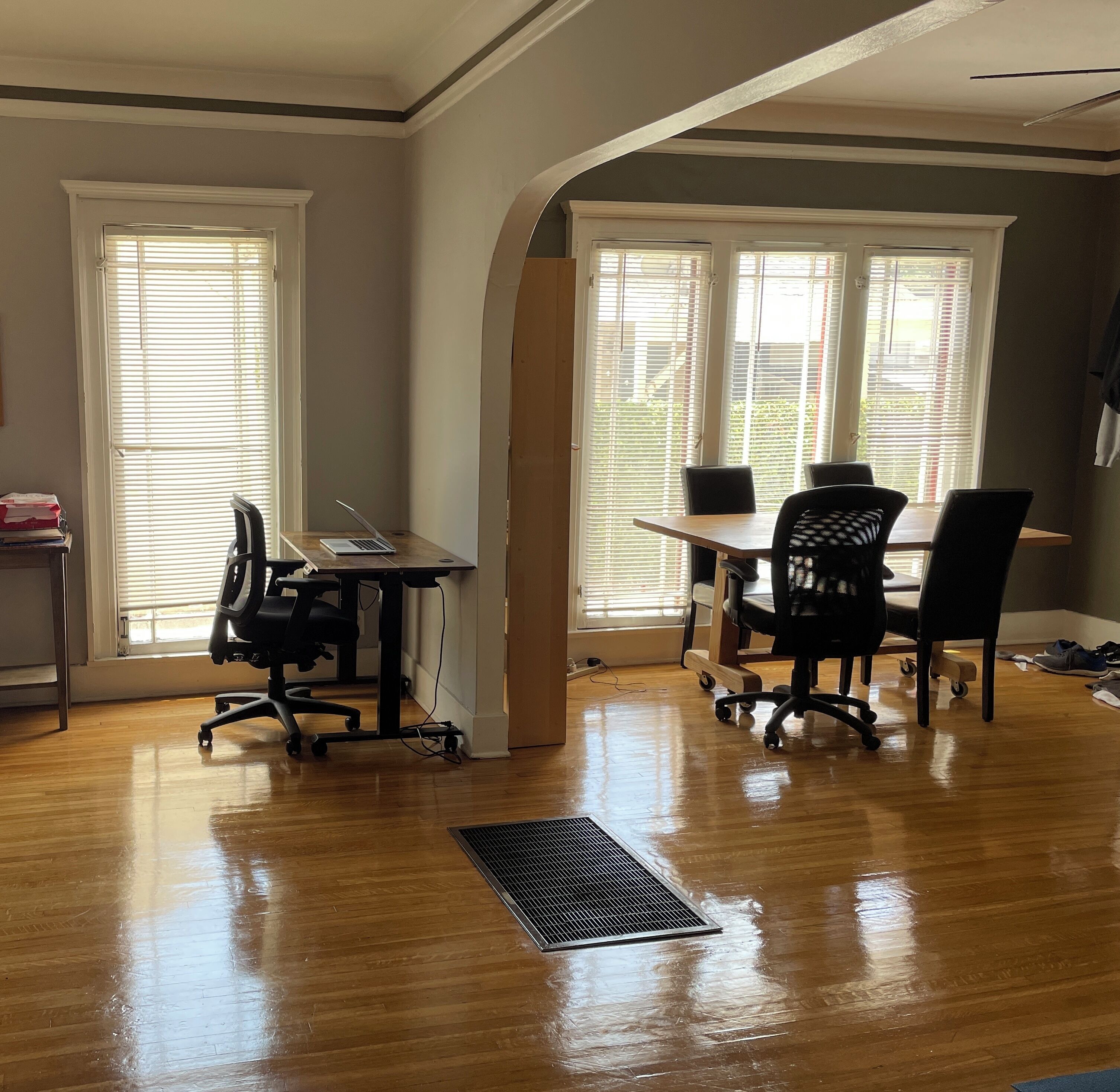 A spacious room with a wooden floor, a desk with a laptop, two chairs, and a large window with blinds.