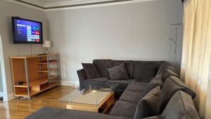 Een moderne woonkamer met een hoekbank, een glazen salontafel en een flatscreen televisie aan de muur.