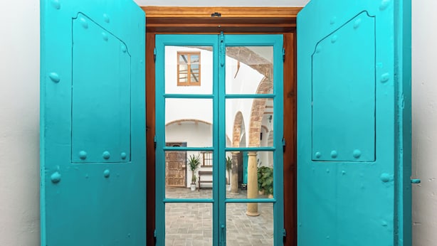 Une porte turquoise avec une fenêtre, donnant sur une cour intérieure où se trouvent un banc et des plantes en pot.