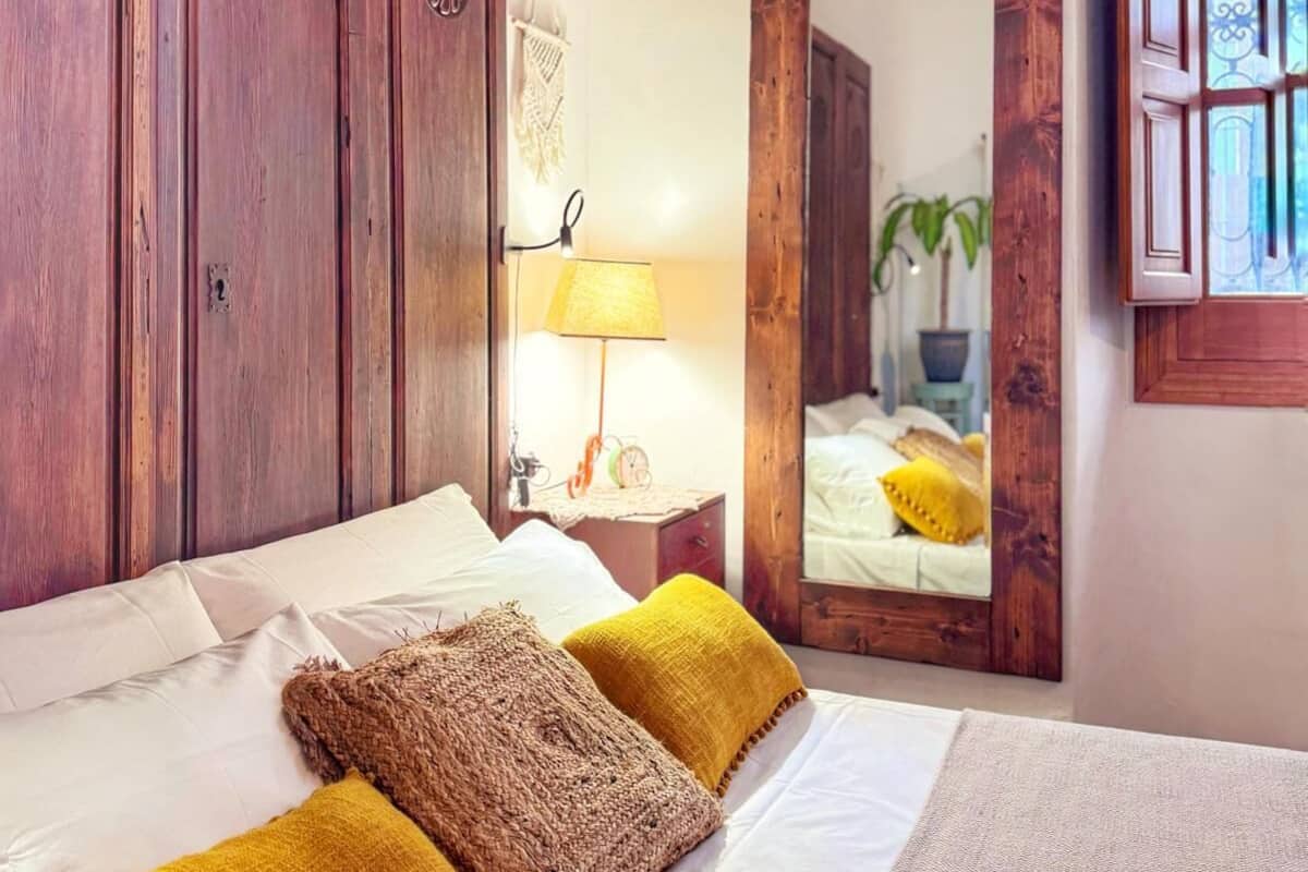 A bedroom with a large bed, wooden wardrobe, a mirror, and a nightstand with a lamp.