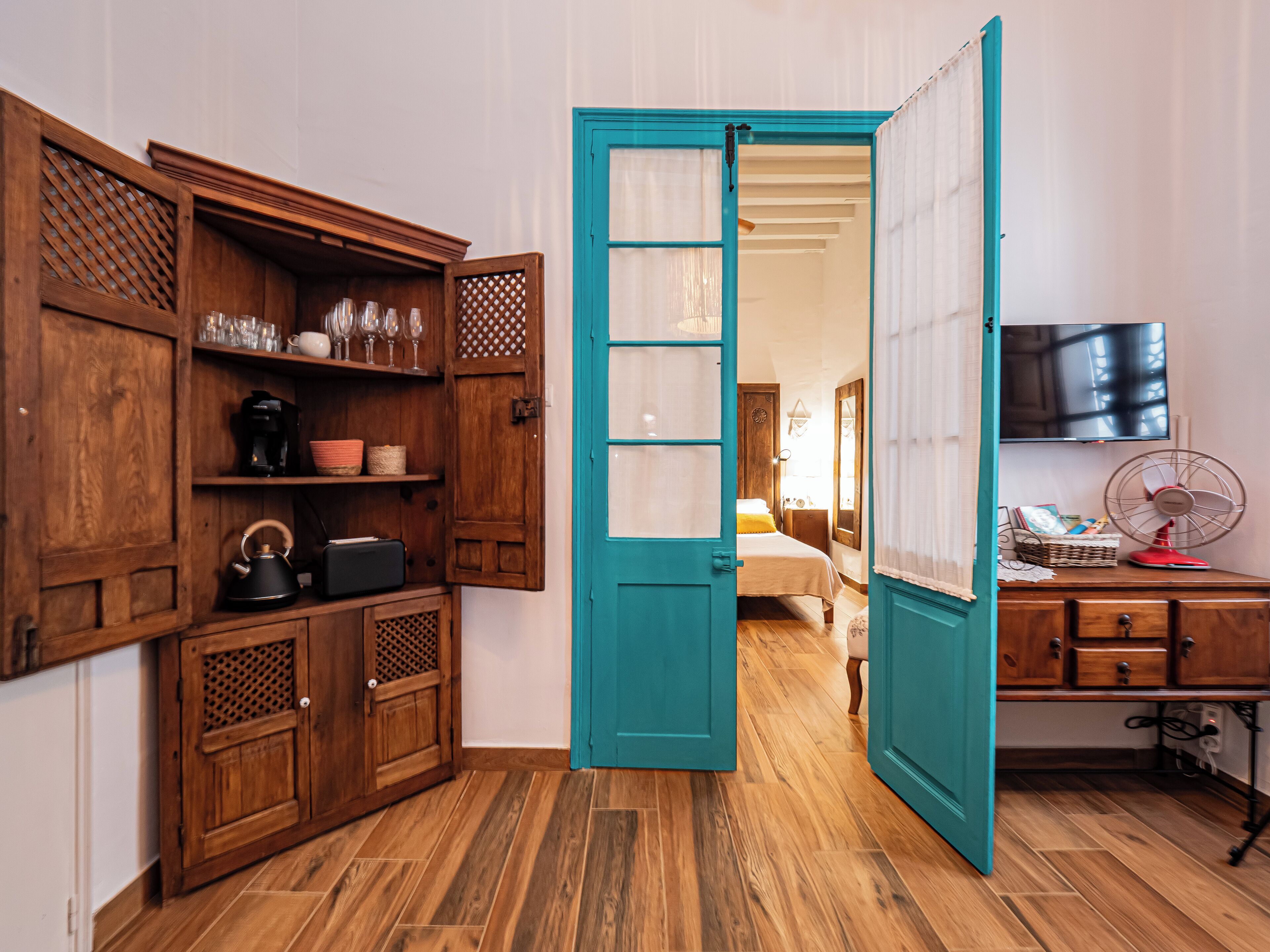 A room with wooden cabinets, a teal door, a bed, a TV, and a fan.