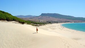 Une plage où l’on voit une personne se promener sur le sable, un ciel dégagé et une ville en arrière-plan.
