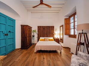 A bedroom with a wooden floor, a bed with a white bedspread, a wooden wardrobe, a blue door, and a window with wooden shutters. - El Conventillo de San Roque (San Roque)