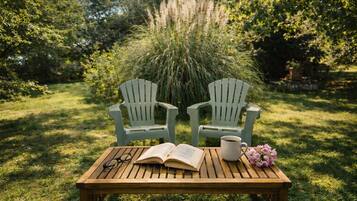 Une table en bois sur laquelle repose un livre ouvert, une paire de lunettes, une tasse et un bouquet de fleurs, le tout dans un jardin agrémenté de deux chaises Adirondack.