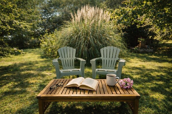 A wooden table with an open book, a pair of glasses, a mug, and a bouquet of flowers in a garden setting with two Adirondack chairs.