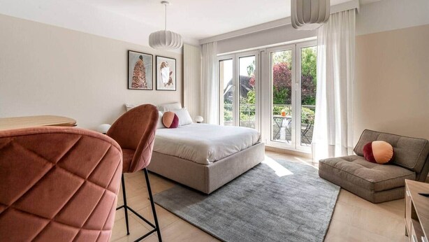 A modern bedroom with a bed, a desk, a chair, and a sofa. There are large windows and a balcony visible. - Studio 4* with balcony and parking in Bel Air (Luxembourg)