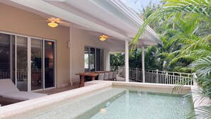 Una piscina con borde de azulejos, un patio techado con mesa y sillas, y vistas a palmeras.