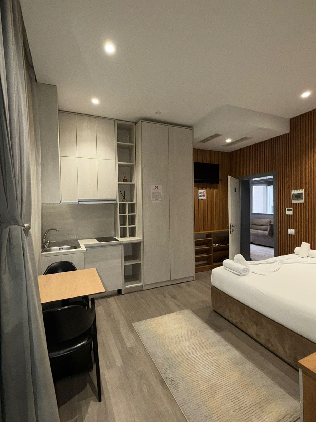 A modern hotel room with a kitchenette, a bed with towels, a desk, and a chair.