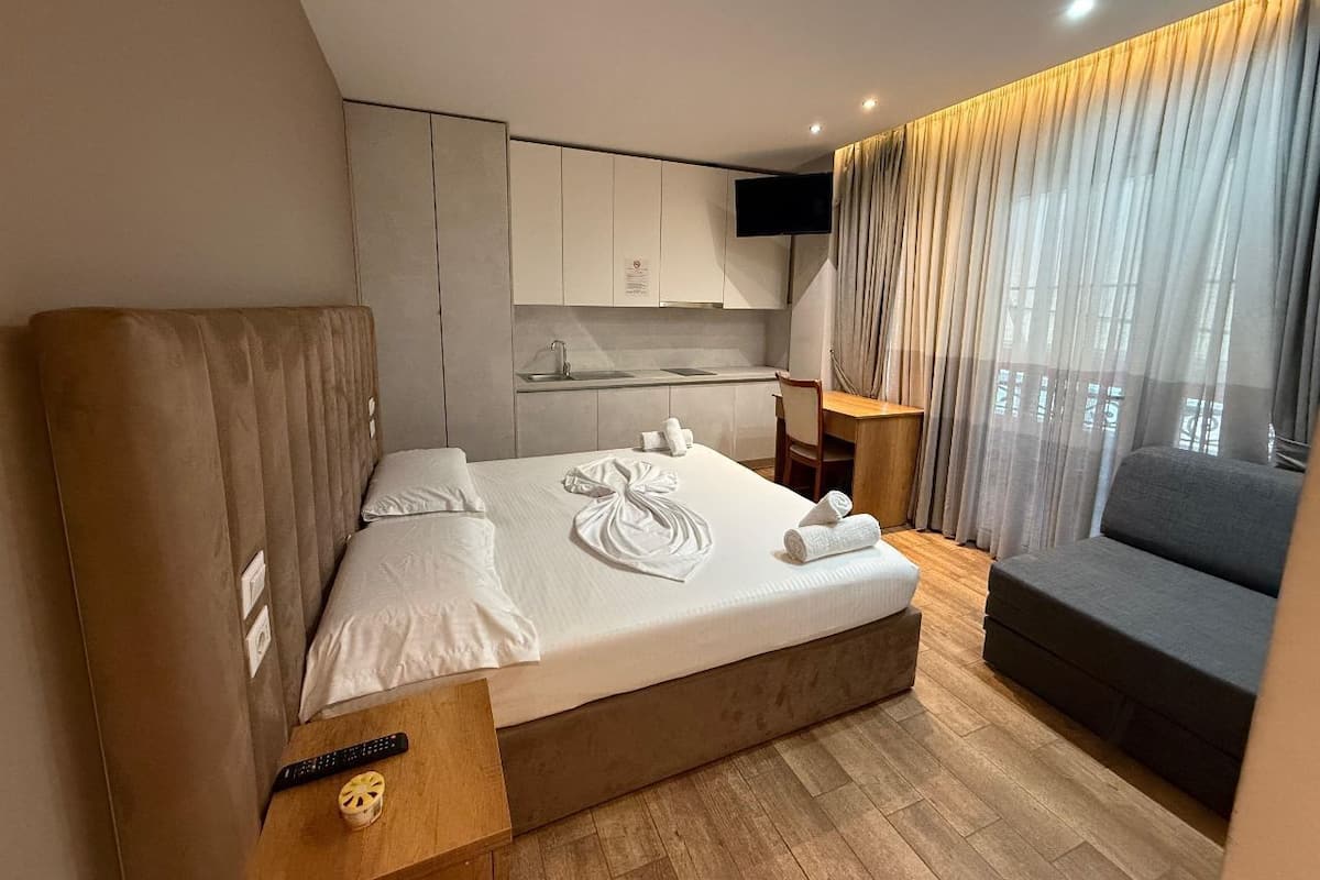 A modern hotel room with a large bed, a small desk, a sofa, a TV, and a kitchen area.