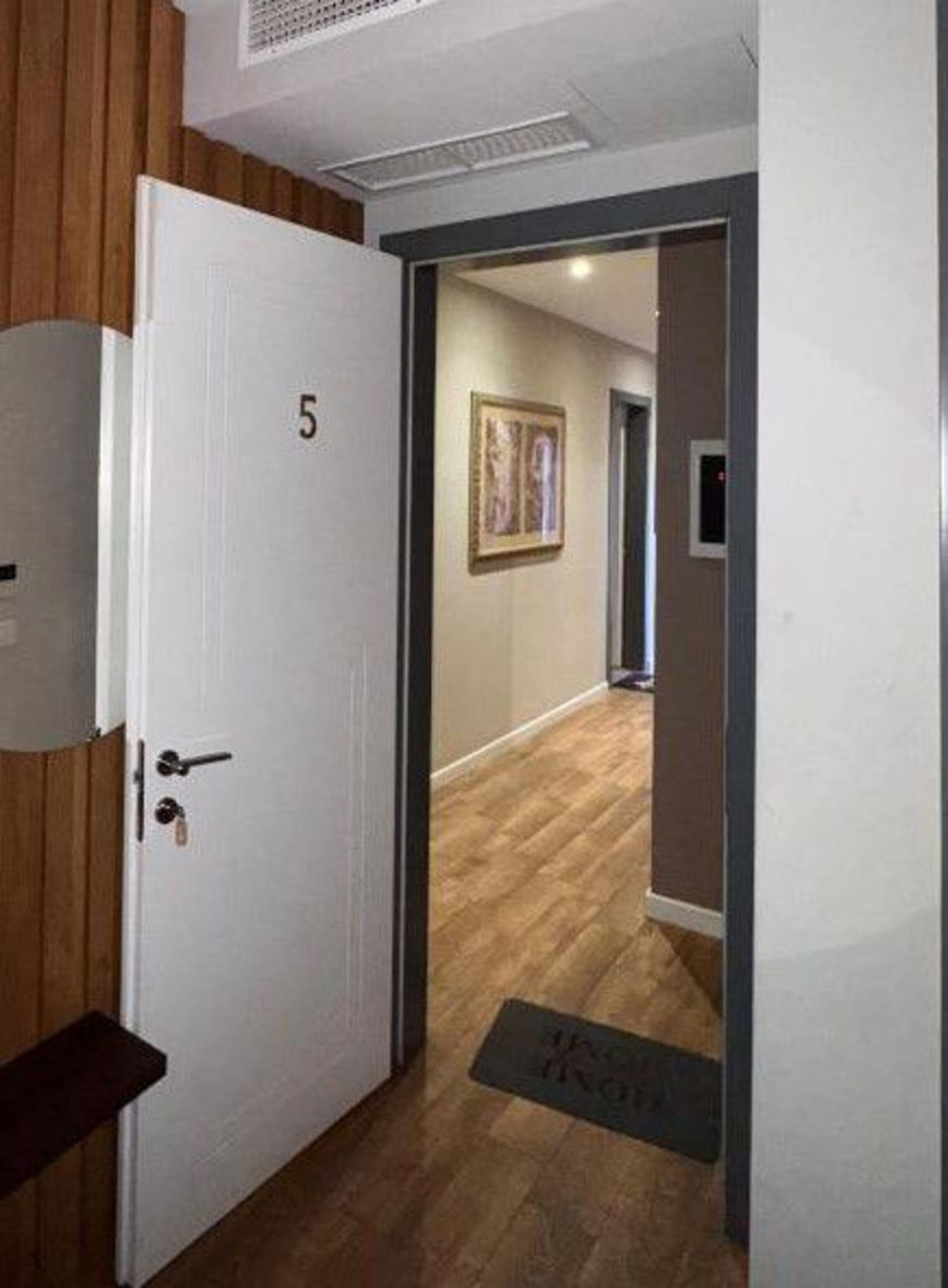 Una habitación de hotel con una puerta de madera, el número 5 en la puerta, un felpudo y un pasillo con suelo de madera y un cuadro enmarcado en la pared.