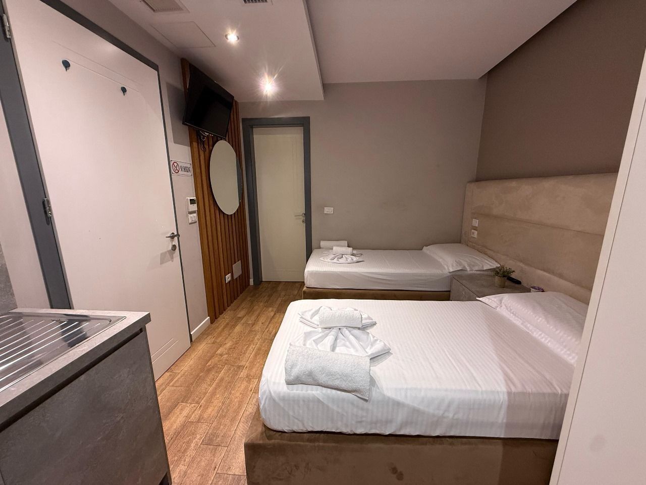 Habitación de hotel con dos camas, un televisor, suelo de madera y una puerta con letrero de 'No Fumar'.