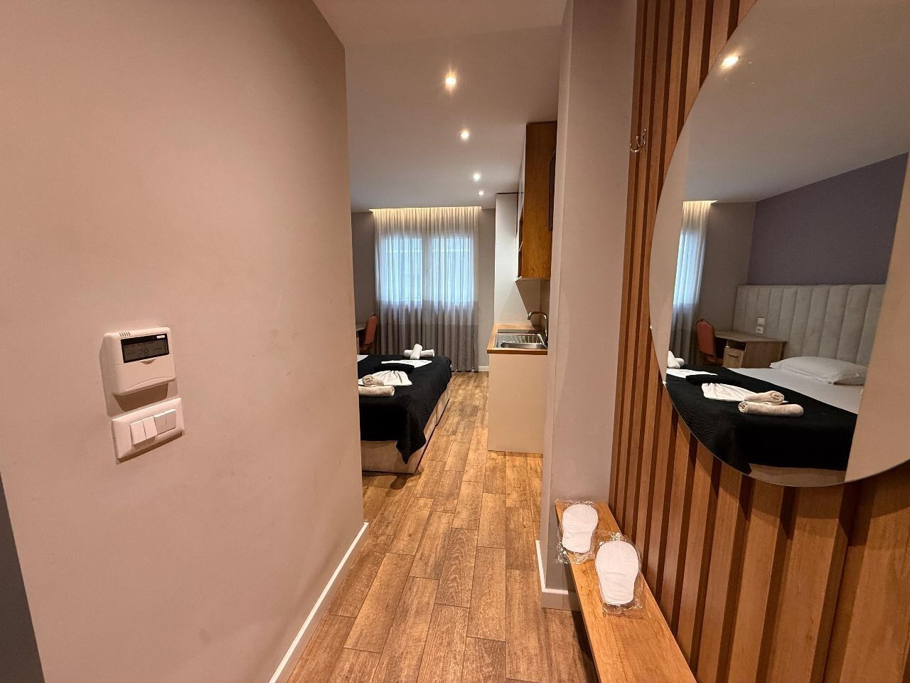 A hotel room with a wooden floor, a bed with a black bedspread, a small desk, a mirror, and a wall-mounted thermostat.