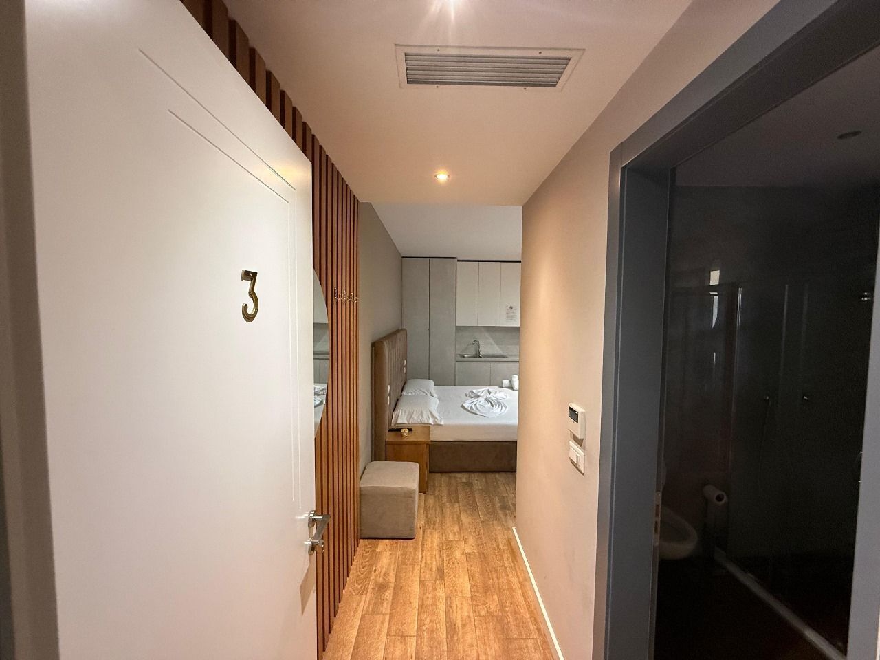 A narrow hallway leading to a bedroom with a bed, a small bench, and a visible bathroom door.