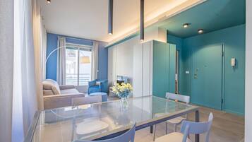 A modern dining area with a glass table and white chairs, a sofa, and a TV in the background.