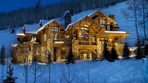 A large, multi-story log cabin with a stone foundation, surrounded by snow-covered trees and a mountain backdrop. - Ski-In Elegance in Arrowhead | High Road (Edwards)