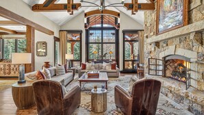 A spacious living room with a stone fireplace, large windows, and a high ceiling with exposed beams. - Ski-In Elegance in Arrowhead | High Road (Edwards)
