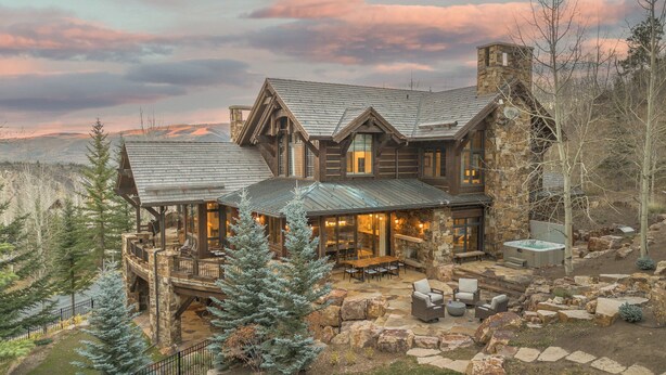 A large log cabin with a stone chimney, a deck with outdoor seating, and a hot tub. - Ski-In Elegance in Arrowhead | High Road (Edwards)