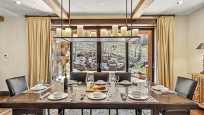 A dining area with a large table set for six, surrounded by chairs, with a view of a rocky outdoor landscape through large windows. - Ski-In Elegance in Arrowhead | High Road (Edwards)