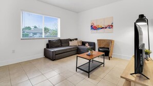Ein modernes Wohnzimmer mit einem grauen Ecksofa, einem Holztisch, einer schwarzen Stehlampe und einem Flachbildfernseher auf einem hölzernen Fuß.