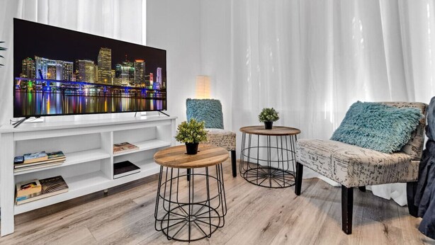 Ein modernes Wohnzimmer mit einem großen Flachbildfernseher, zwei runden Tischen mit Topfpflanzen, einem Sofa mit blauem Zierkissen und einer weißen Unterhaltungsecke mit Büchern.