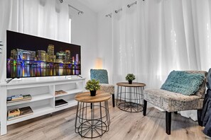 A modern living room with a large flat-screen TV, two round tables with potted plants, a sofa with a blue throw pillow, and a white entertainment unit with books. - °Wonderful 6ppl Apt 5 min walking Design District° (Miami)