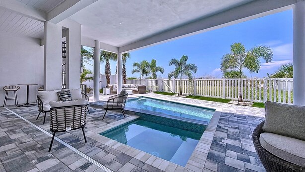 A covered outdoor area with a swimming pool, seating, and a view of palm trees. - Beach Two: Style Meets the Shore w/ Pool and Spa (Treasure Island)
