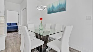 Uma área de refeições moderna com mesa de vidro, cadeiras brancas e um quadro azul na parede.