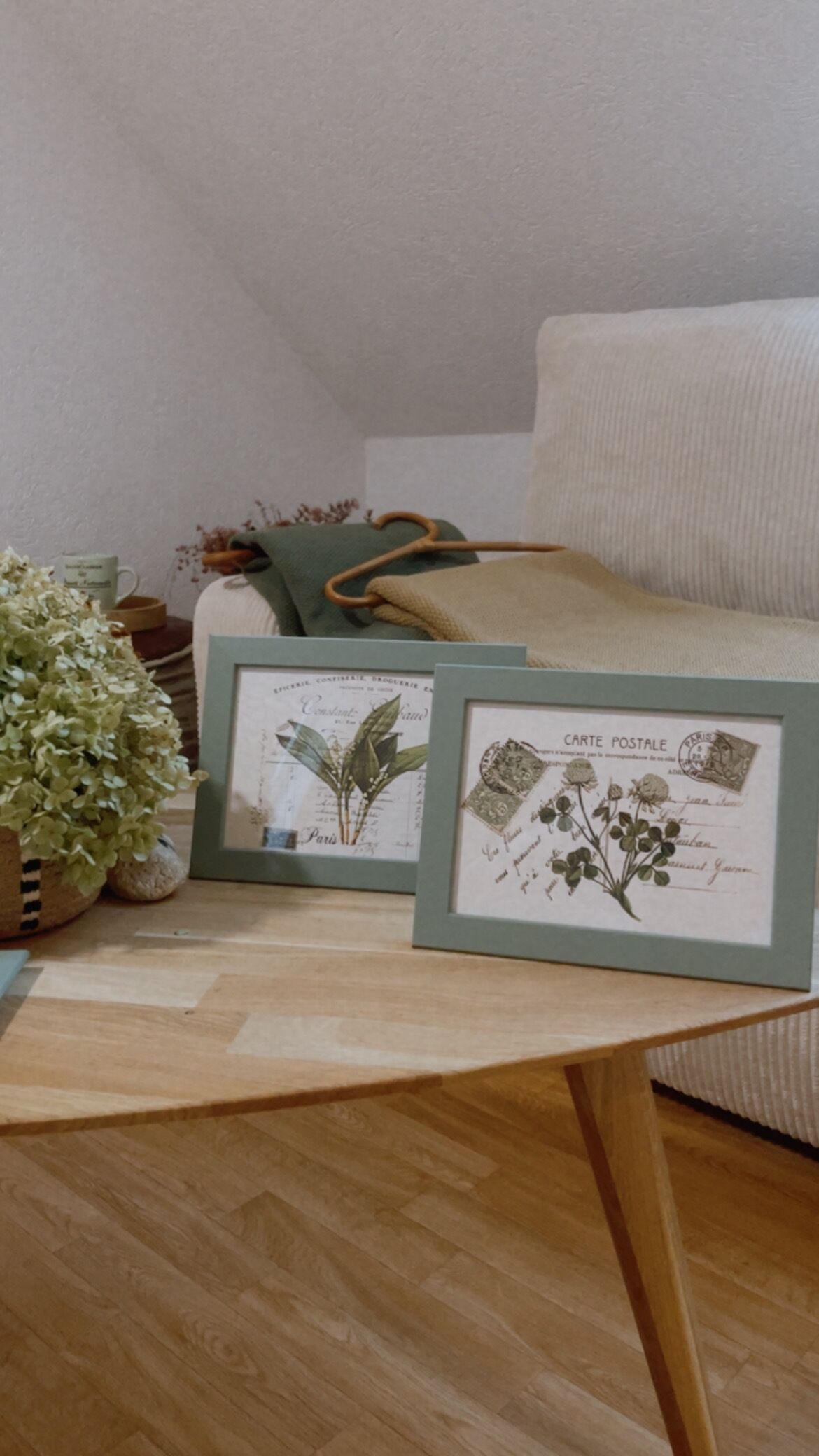 Une pièce avec un plancher en bois, un lit recouvert d’une couverture verte, une estampe botanique encadrée et une petite plante en pot.