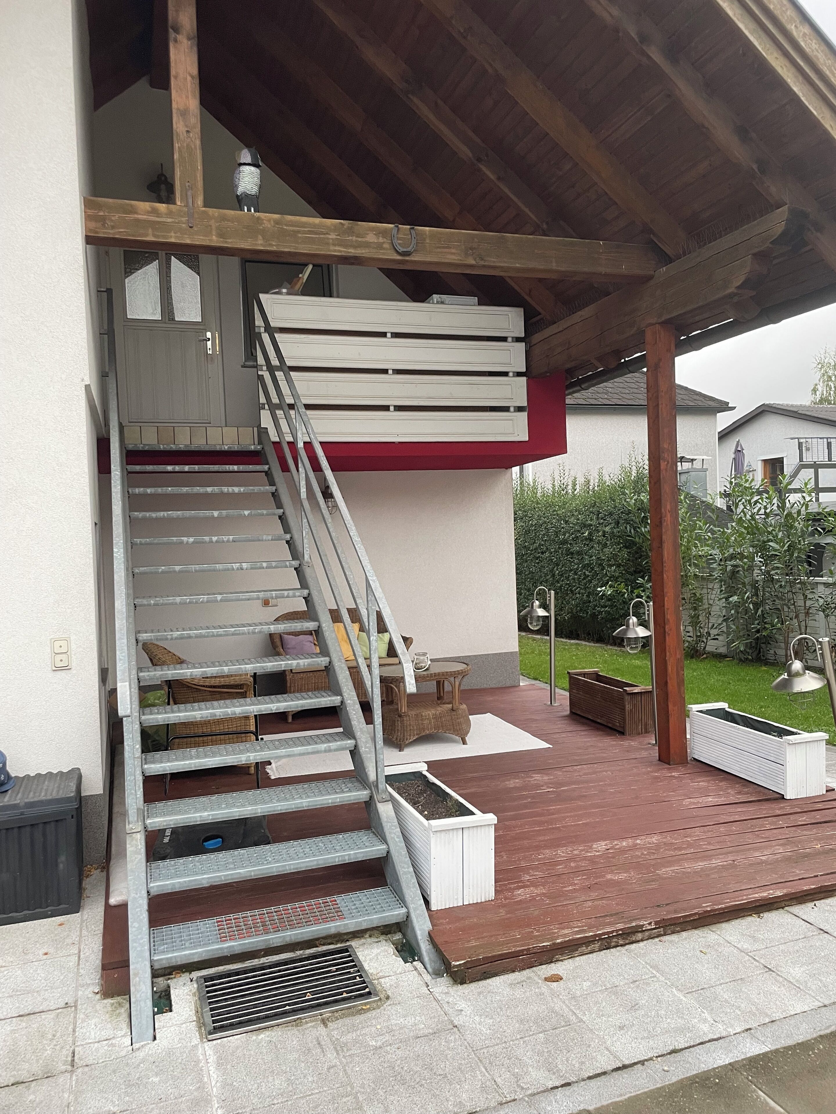 A covered outdoor area with a wooden staircase leading to a white door, a small table with chairs, and a planter box.