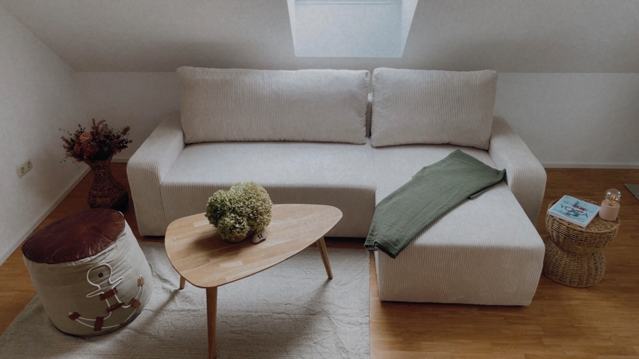 A modern living room with a sofa, a small wooden table, a decorative stool, and a wicker side table with a book and a candle.