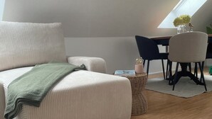 Une chambre à coucher confortable, aménagée en style « loft », avec une grande fenêtre, un canapé confortable, une table à manger et une petite table d’appoint agrémentée d’un vase et d’une bougie.
