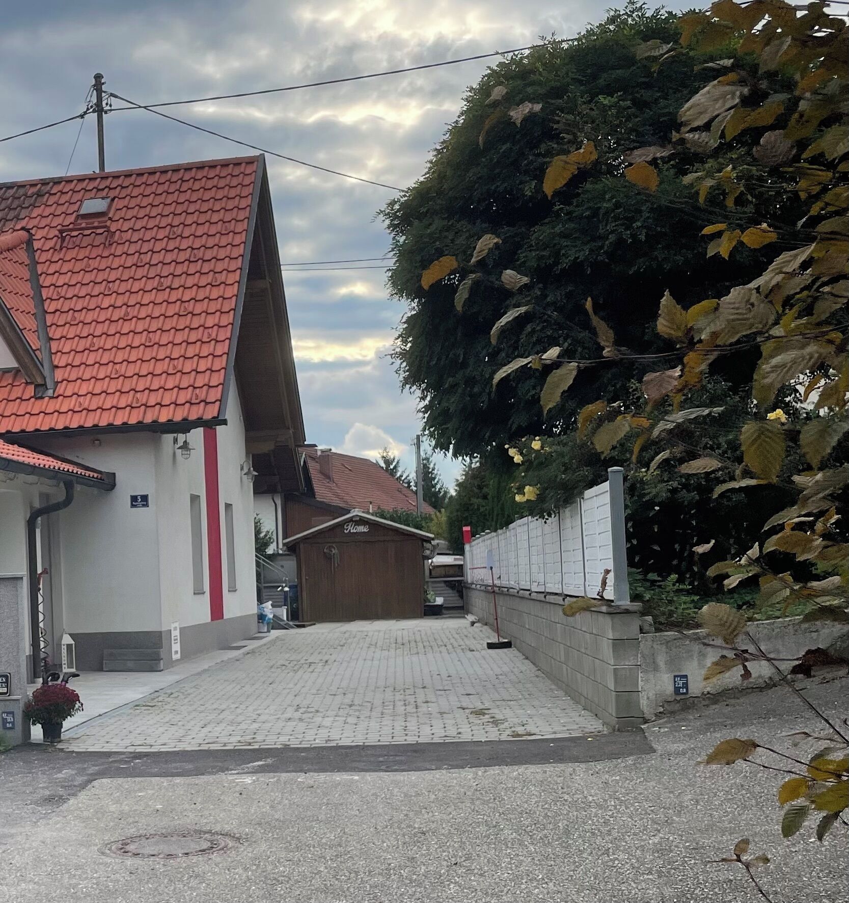 Una strada residenziale con una casa a tetto in tegole rosse, un cancello in legno con la scritta 'Home' e un vialetto asfaltato.