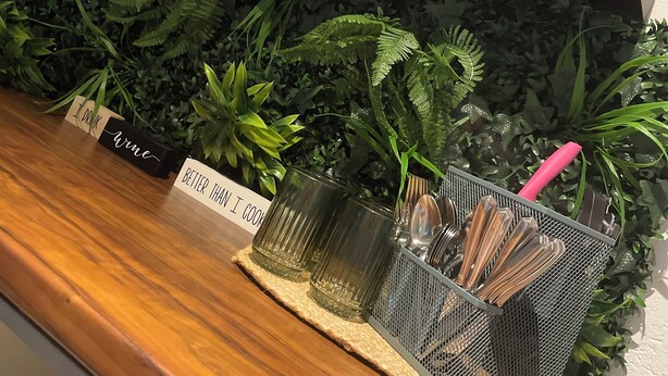 Un comptoir en bois surmonté d’un récipient en treillis métallique contenant des ustensiles, une étagère avec une plante et une pancarte sur laquelle on peut lire : « Je bois du vin. »