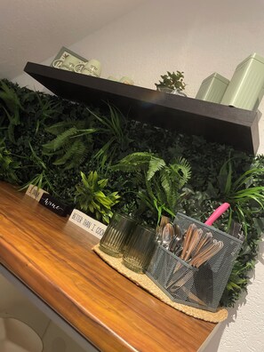 A wooden counter with a metal mesh container holding utensils, a shelf with a plant, and a sign that reads "I drink wine." - Quaint 1-bedroom apartment in Bad Schallerbach (Bad Schallerbach)