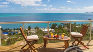 Een balkon met twee houten stoelen, een klein rond tafeltje met fruit en drankjes, en uitzicht op de oceaan en het resort.
