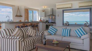 Een woonkamer met gestreepte banken, een bijzettafeltje met een fles en glazen, en uitzicht op de oceaan door grote ramen.