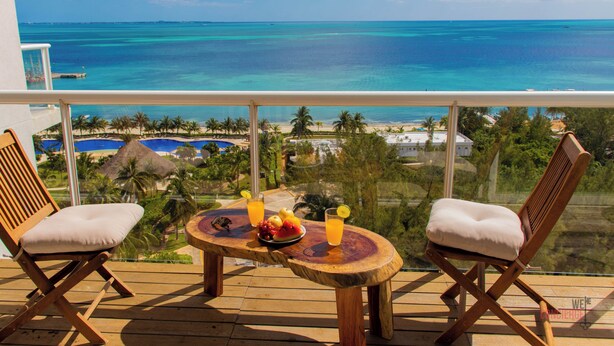 Een balkon met twee houten stoelen, een klein rond tafeltje met fruit en drankjes, en uitzicht op de oceaan en het resort.