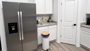 A modern kitchen with white cabinets, a stainless steel refrigerator, a wooden stool, and a trash bin. - Wonderful 3-bedroom house with AC in Fort Myers (Fort Myers)