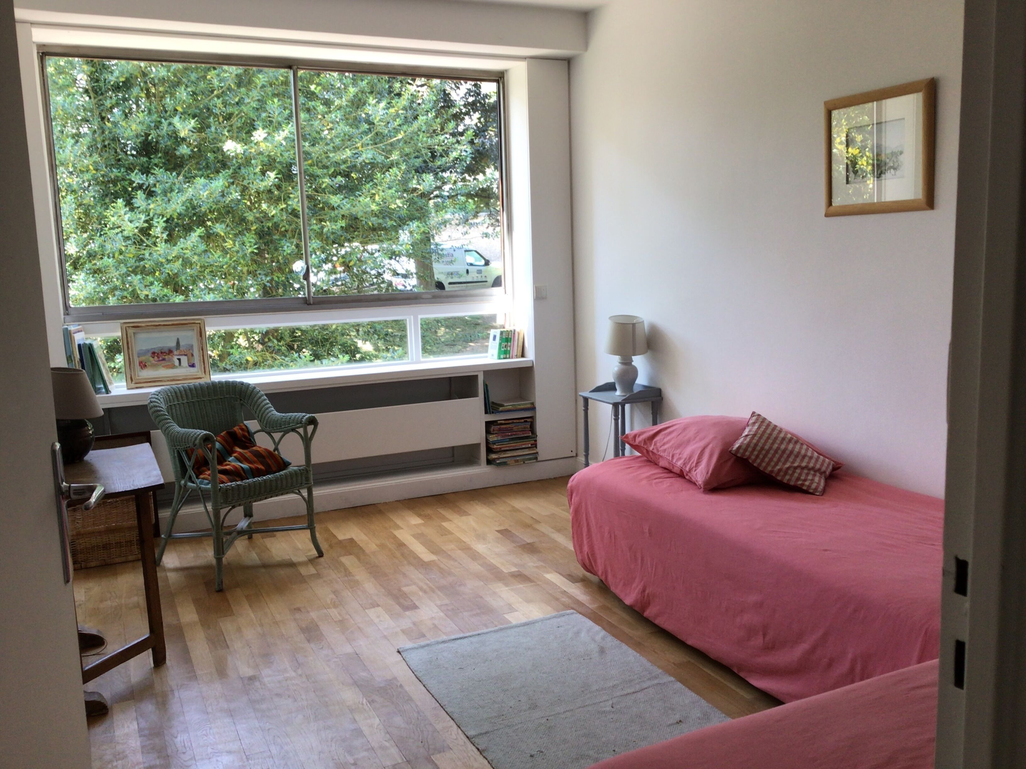 A room with a large window, a bed with pink bedding, a chair, a small table with a lamp, and a bookshelf.