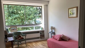 A room with a large window, a bed with pink bedding, a chair, a small table with a lamp, and a bookshelf. - Spacious, bright apartment near Paris (Louveciennes)