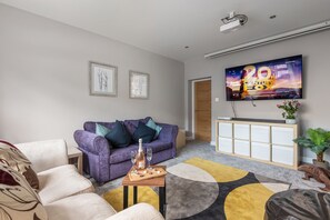 A modern living room with a purple sofa, a wooden side table with a bottle and glasses, a large flat-screen TV, and a decorative rug. - Newlands (Yelverton)