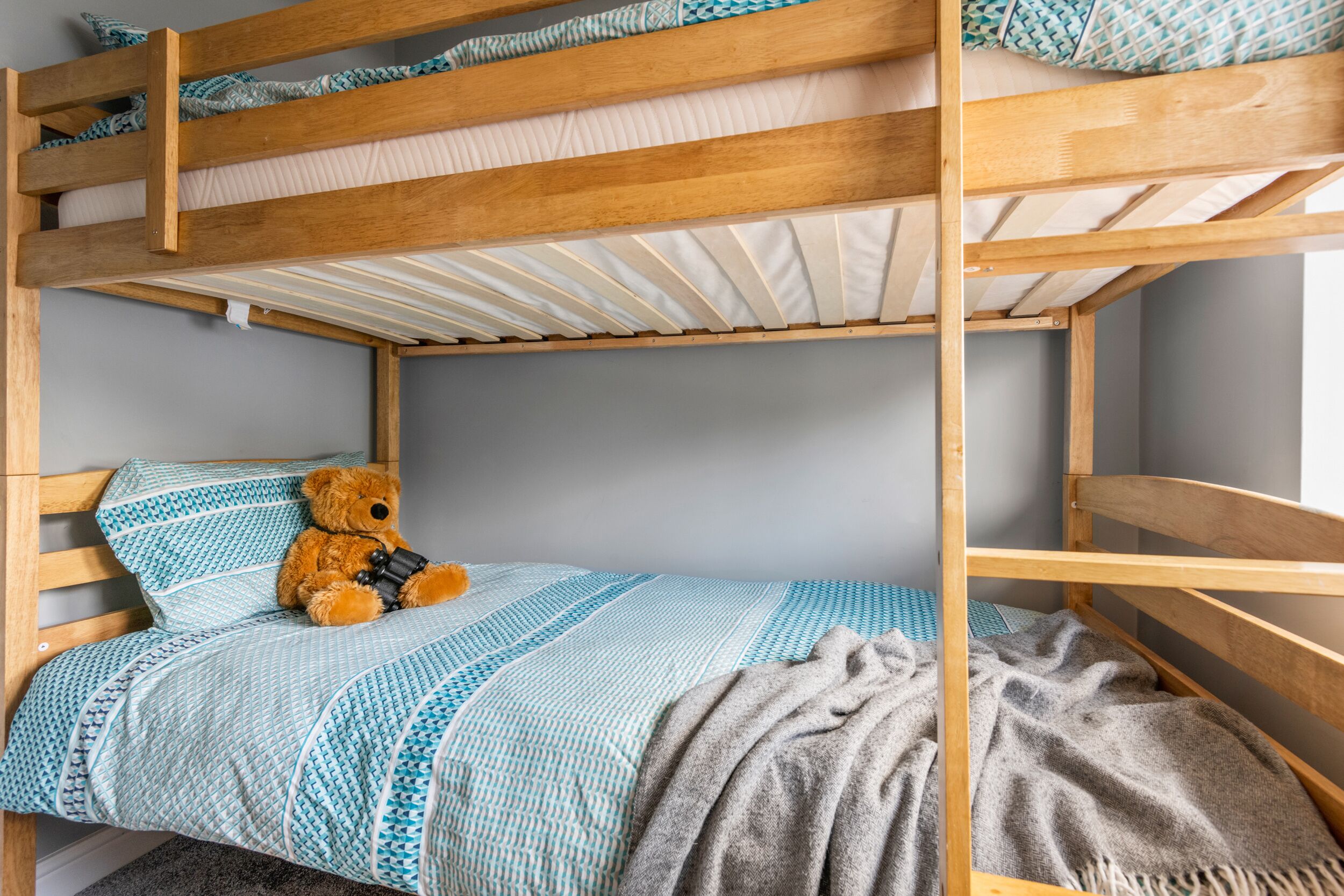 A bunk bed with a teddy bear on the lower bunk, a patterned blanket, and a grey throw blanket.
