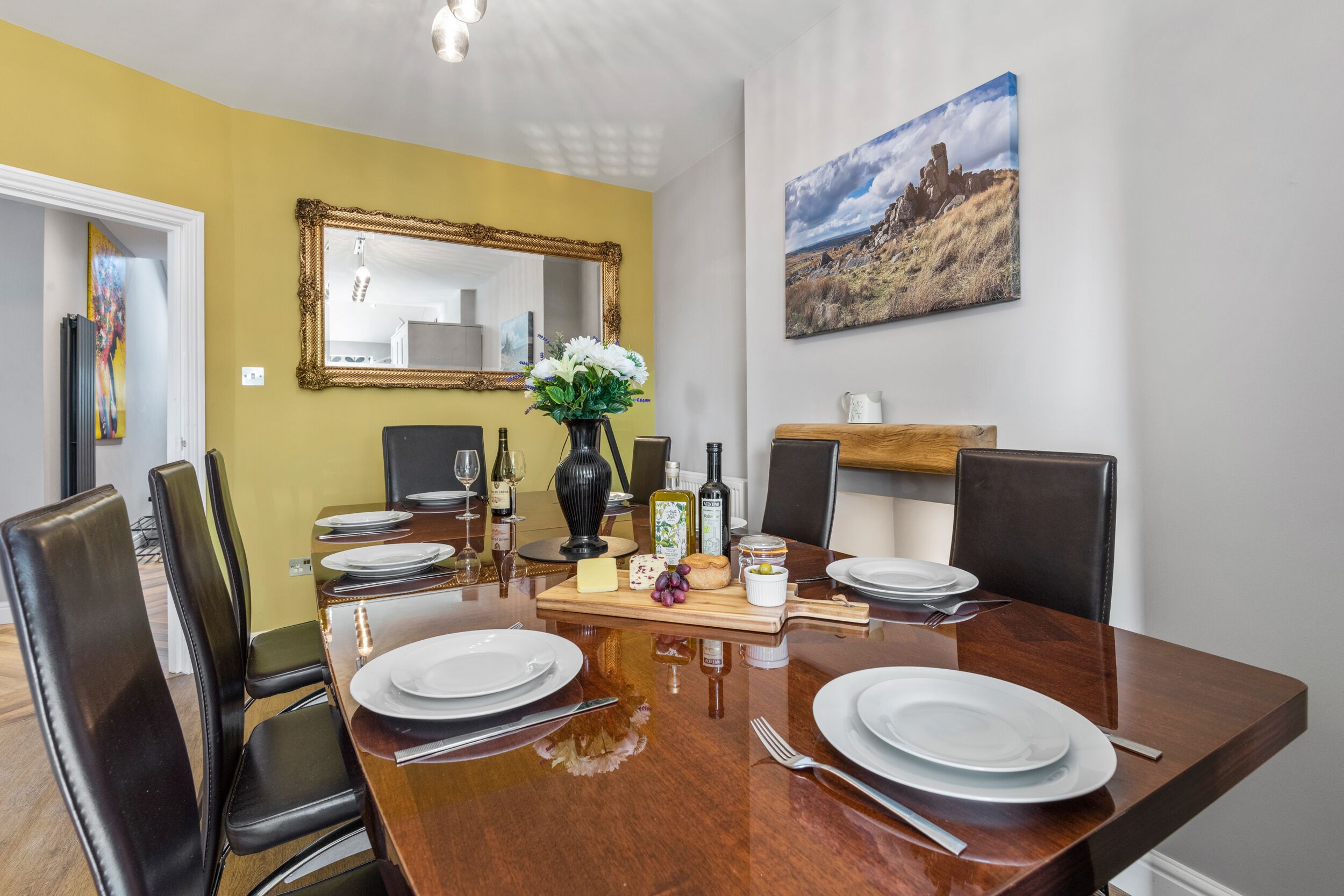 Comedor con una mesa de madera dispuesta para cuatro personas, rodeada de sillas negras. En las paredes, un espejo y un cuadro.
