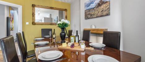 Comedor con una mesa de madera dispuesta para cuatro personas, rodeada de sillas negras. En las paredes, un espejo y un cuadro.