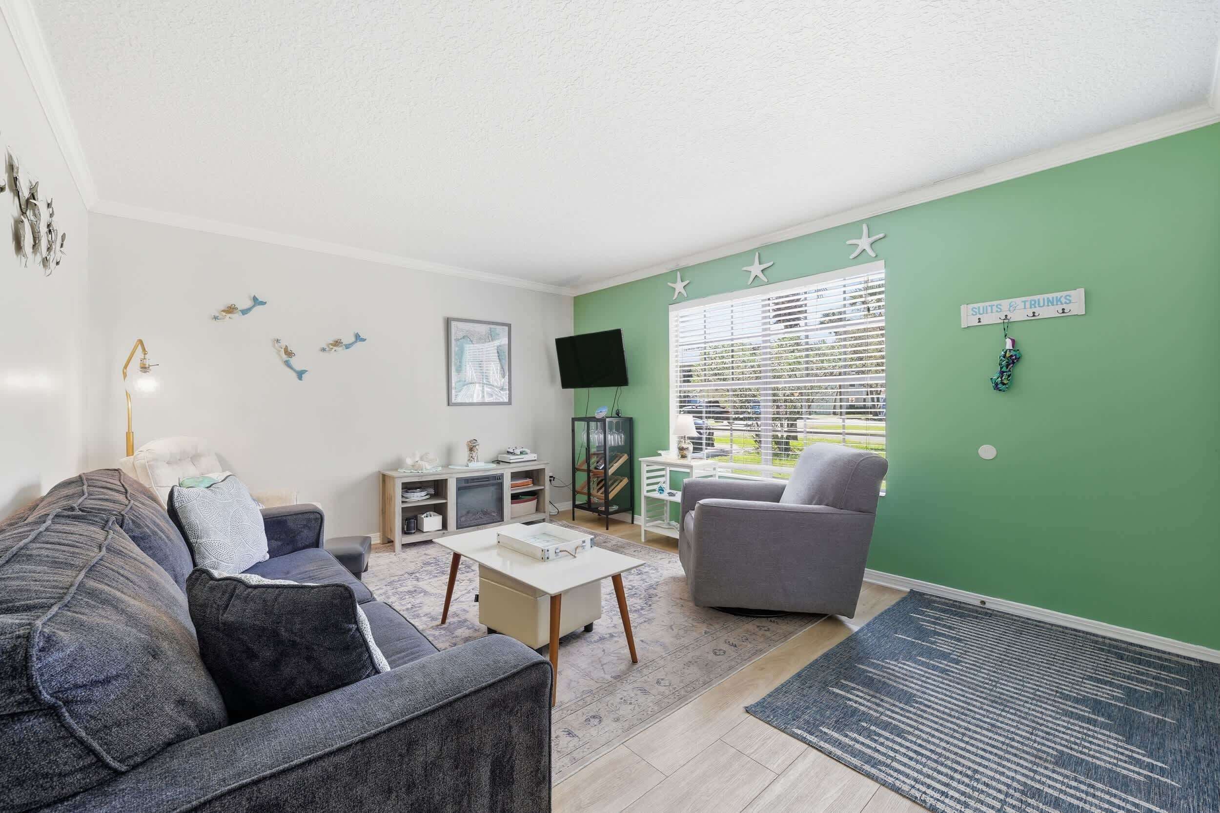 A living room with a green accent wall, a grey sofa, a coffee table, a TV, and a window with blinds.