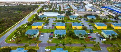 An aerial view of a coastal residential area with multiple two-story yellow buildings, a beachfront promenade, and a large hotel complex.