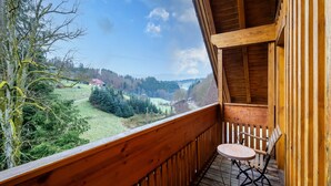 Un balcón de madera con una mesa y una silla, con vista a un valle lleno de árboles y casas.